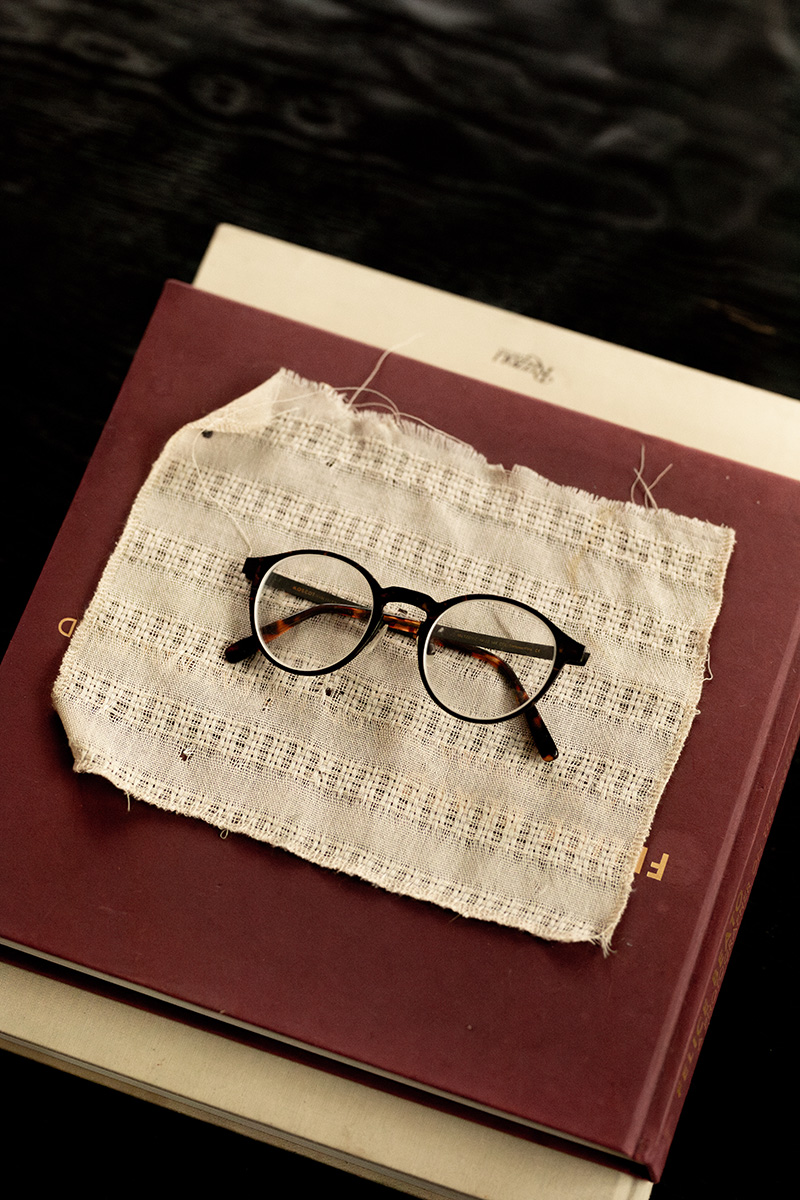 A pair of round eyeglasses is placed on a piece of fabric atop two stacked books with dark covers.