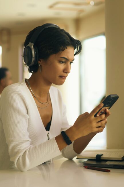 A person wearing headphones and a white cardigan sits at a table, looking at a smartphone. A pen and notebook are on the table.