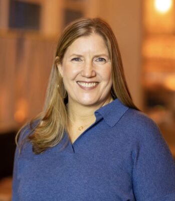 A woman with long, light brown hair is smiling at the camera. She is wearing a blue sweater and standing indoors with a blurred background.