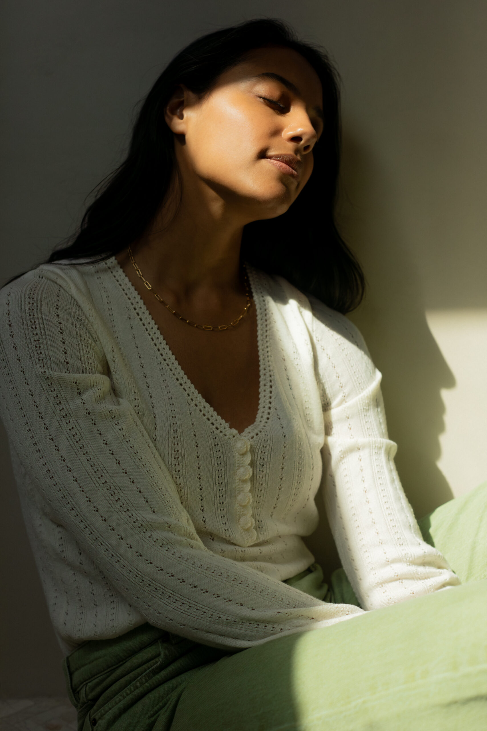 A woman in a white knit top and light green pants sits against a wall, eyes closed, with sunlight partially illuminating her face and casting a shadow behind her.