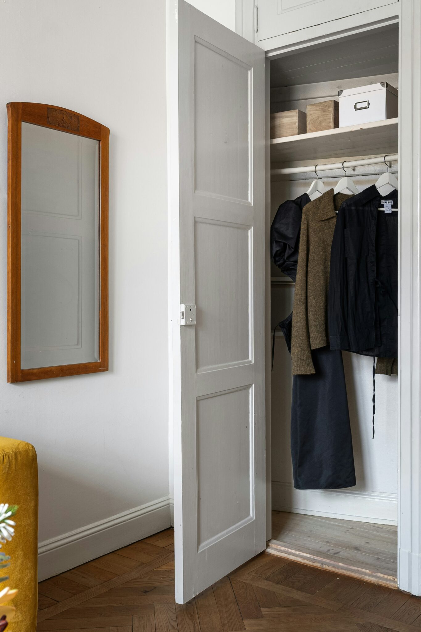 A white closet door is open, revealing clothes hanging and storage boxes on shelves. A wooden-framed mirror is mounted on the adjacent wall.