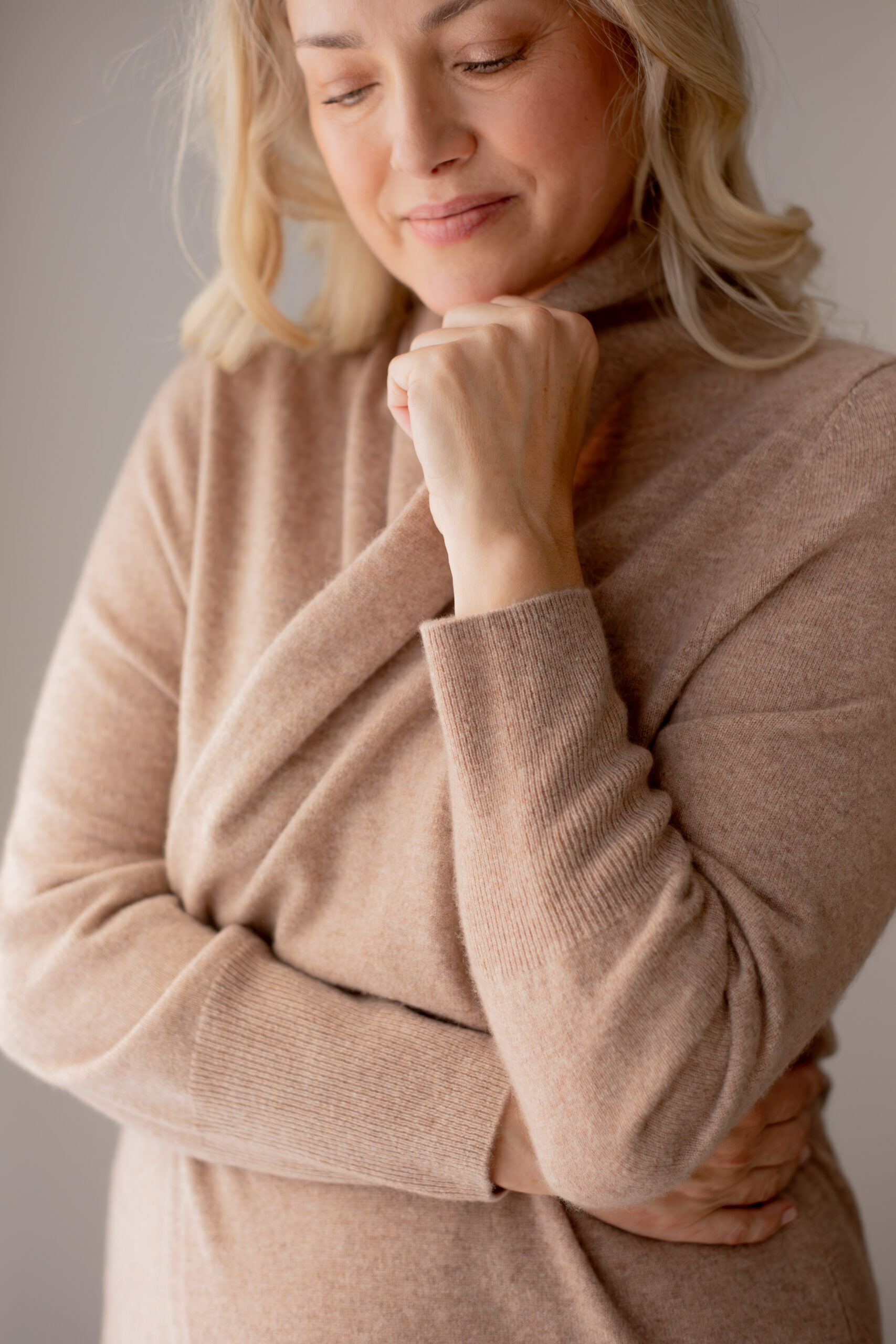 Woman in a beige turtleneck sweater stands with one arm crossed and the other hand resting under her chin, gazing slightly downward with a calm expression.