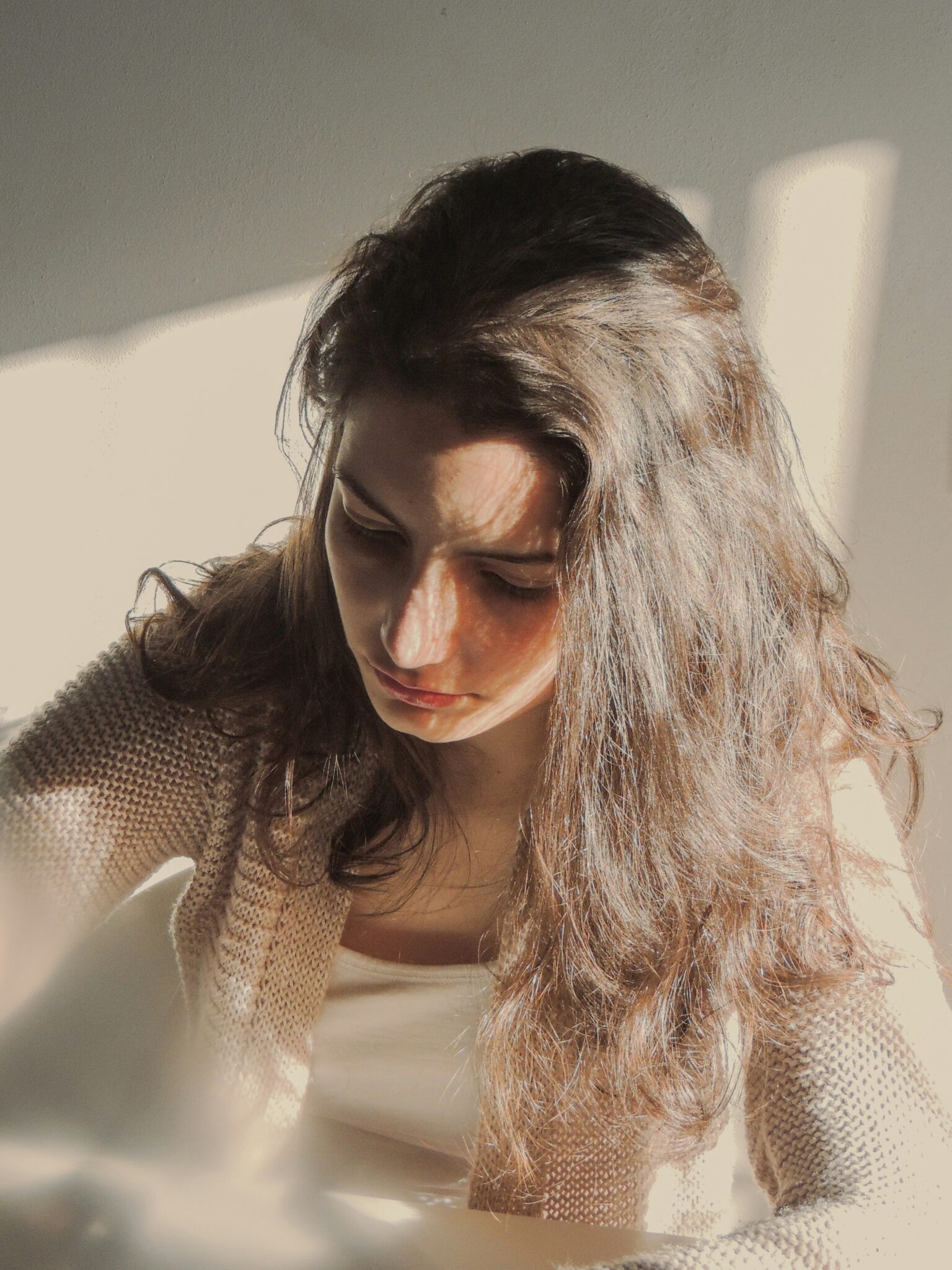 A woman with long brown hair sits in sunlight, looking down with a neutral expression. She wears a beige sweater and a white top.