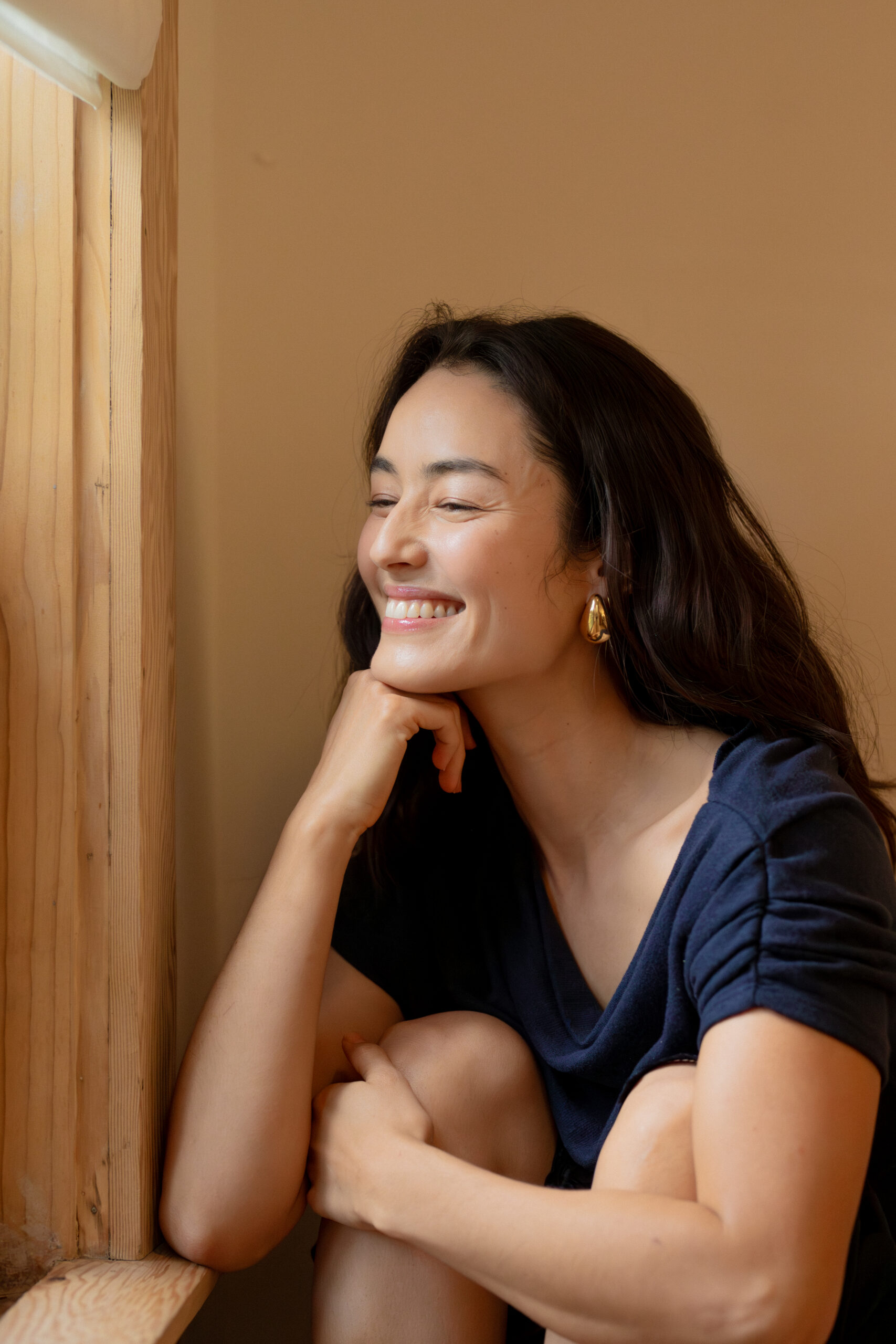 A woman with long dark hair and a navy top sits by a wooden window, resting her chin on her hand and smiling softly.