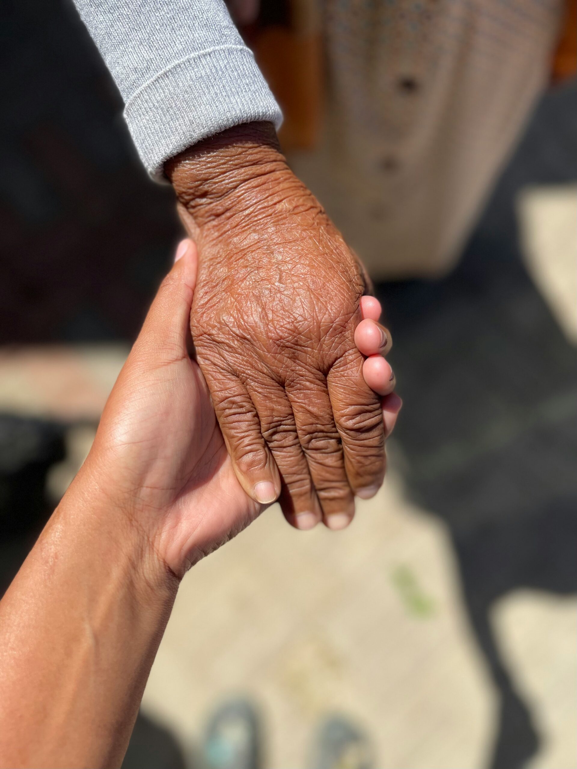 A younger hand gently holds an older hand outdoors, showing a contrast in skin texture and age.