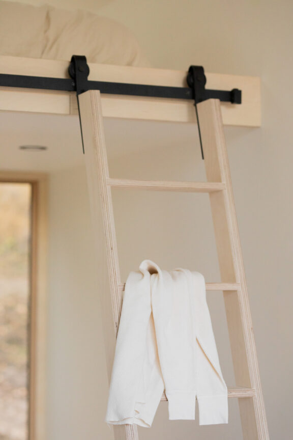 A light wooden ladder leads to a loft bed with bedding. A white sweater is draped over one of the ladder's rungs.