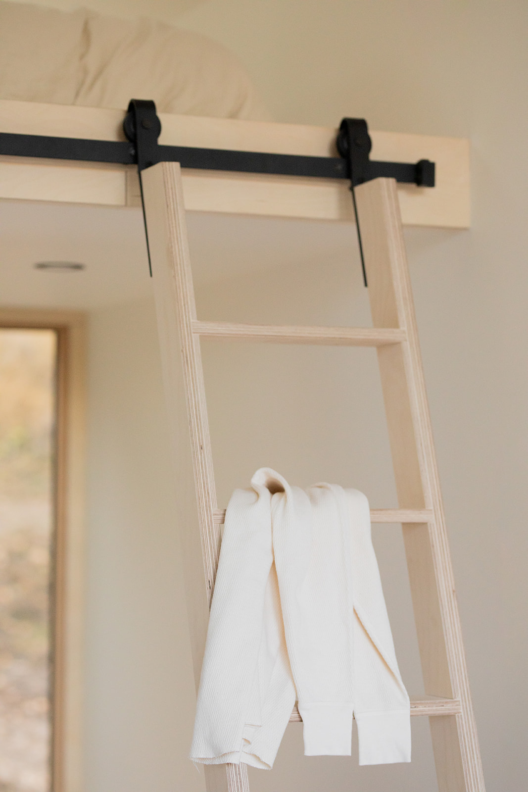 A light wooden ladder leads to a loft bed with bedding. A white sweater is draped over one of the ladder's rungs.