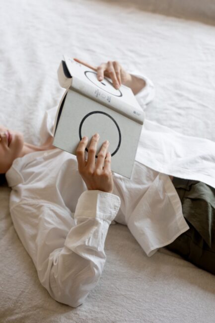 Person lying on a bed, wearing a white shirt and green pants, holding and reading a book with a minimalist cover featuring black circles.