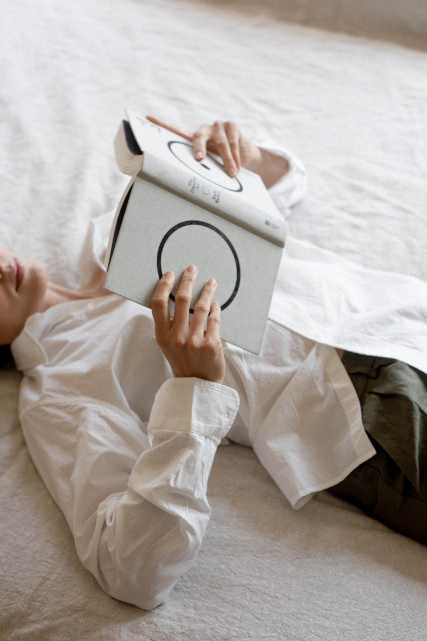 Person lying on a bed, wearing a white shirt and green pants, holding and reading a book with a minimalist cover featuring black circles.
