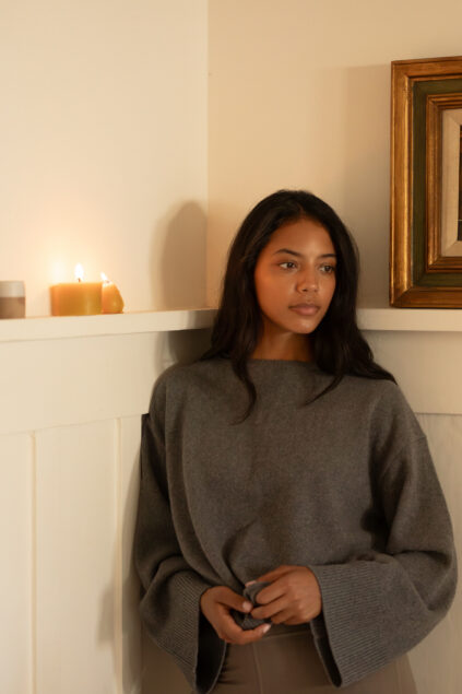 A woman with long dark hair, wearing a loose gray sweater, stands by a white wall next to lit candles and a framed picture.