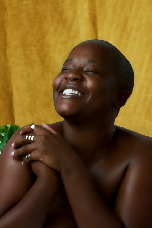 A person with a shaved head smiles with eyes closed, hands clasped near their face, wearing multiple rings, posed against a yellow background.