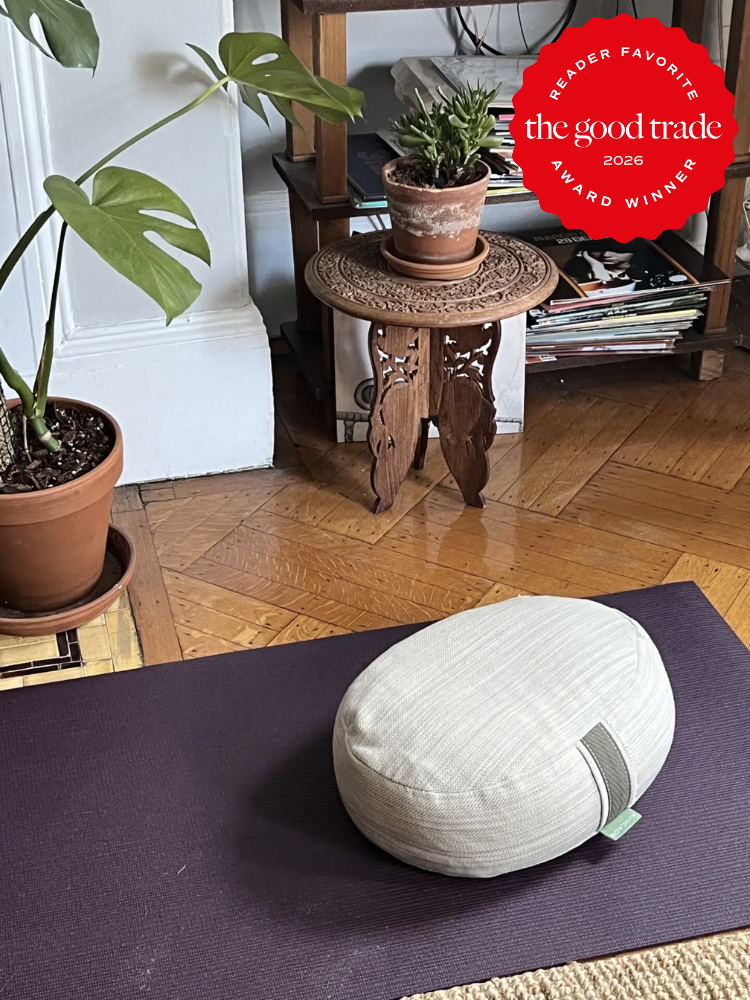 A meditation cushion sits on a yoga mat next to potted plants and a small wooden table with books stacked underneath. A "The Good Trade 2026 Award Winner" badge is in the top right corner.