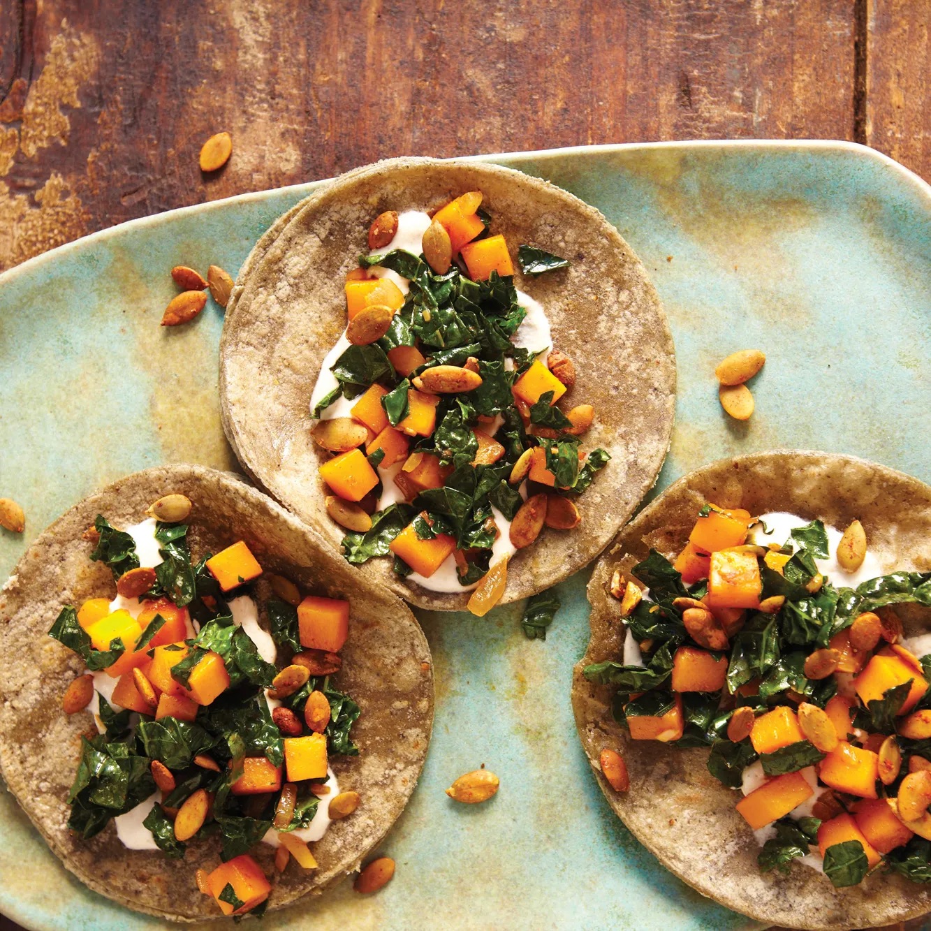 Three corn tortillas topped with a mix of diced squash, sautéed greens, pepitas, and a white sauce on a blue ceramic plate on a wooden surface.