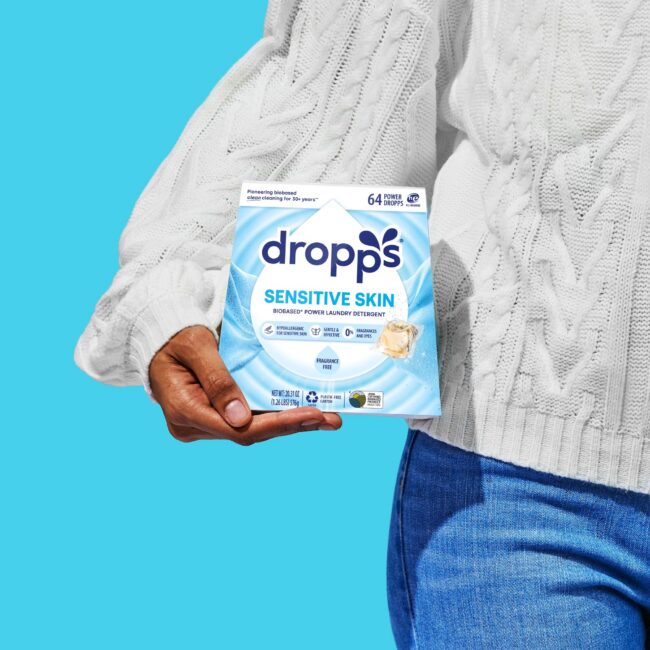 A person in a white sweater and blue jeans holds a box of Dropps Sensitive Skin laundry detergent against a bright blue background.