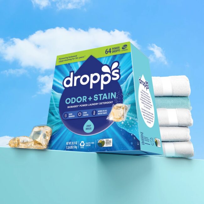 A box of Dropps Odor + Stain laundry detergent pods sits next to a stack of folded white towels with blue stripes against a blue background and sky.