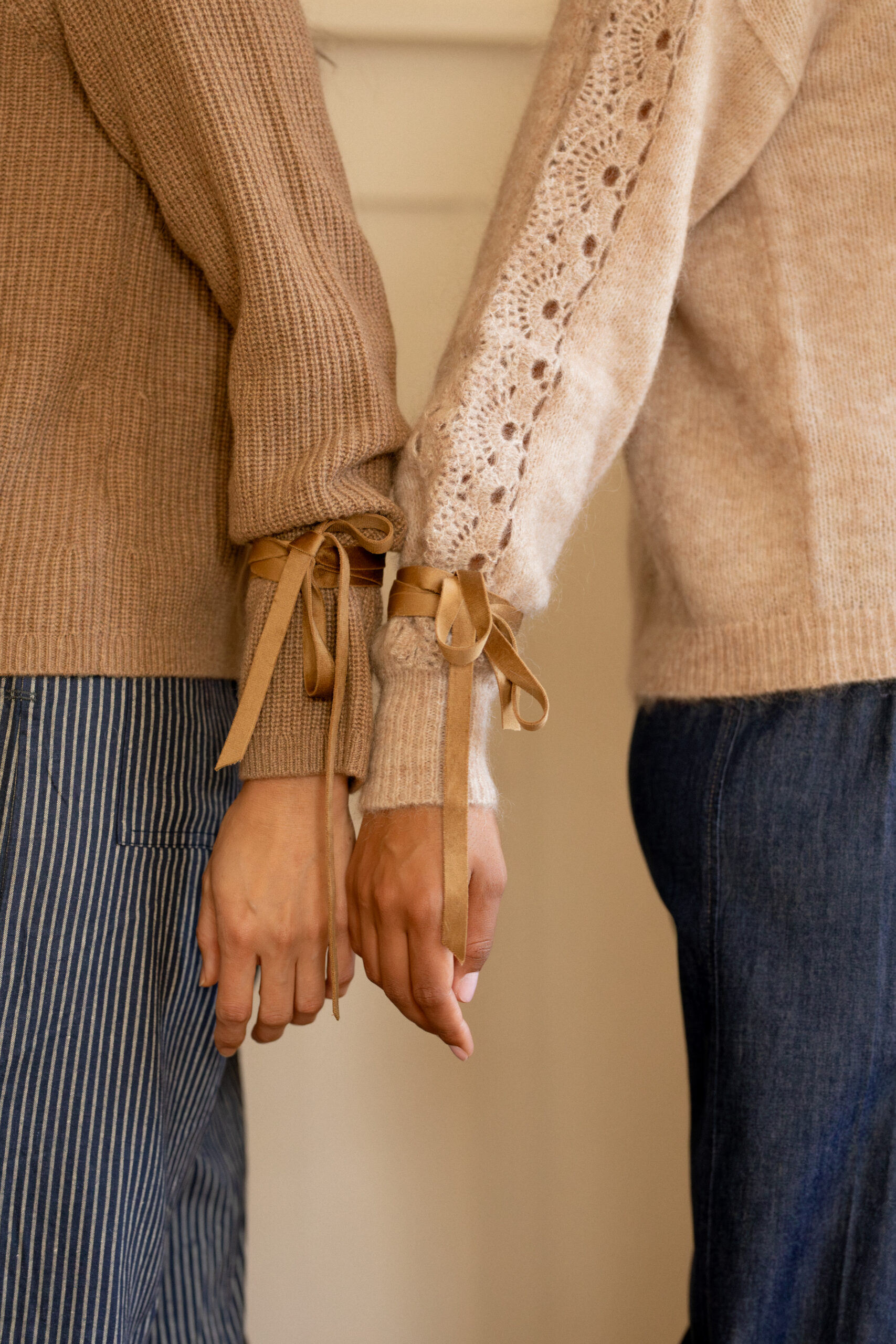 Two people stand side by side, their hands loosely linked. Both wear beige sweaters with ribbon details on the cuffs and blue pants, one striped and the other solid.