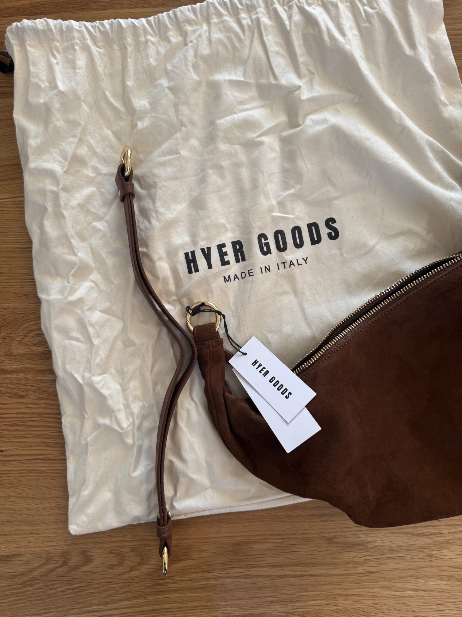 A brown suede bag with a detachable strap and tag is placed on a white dust bag labeled "HYER GOODS MADE IN ITALY" on a wooden surface.