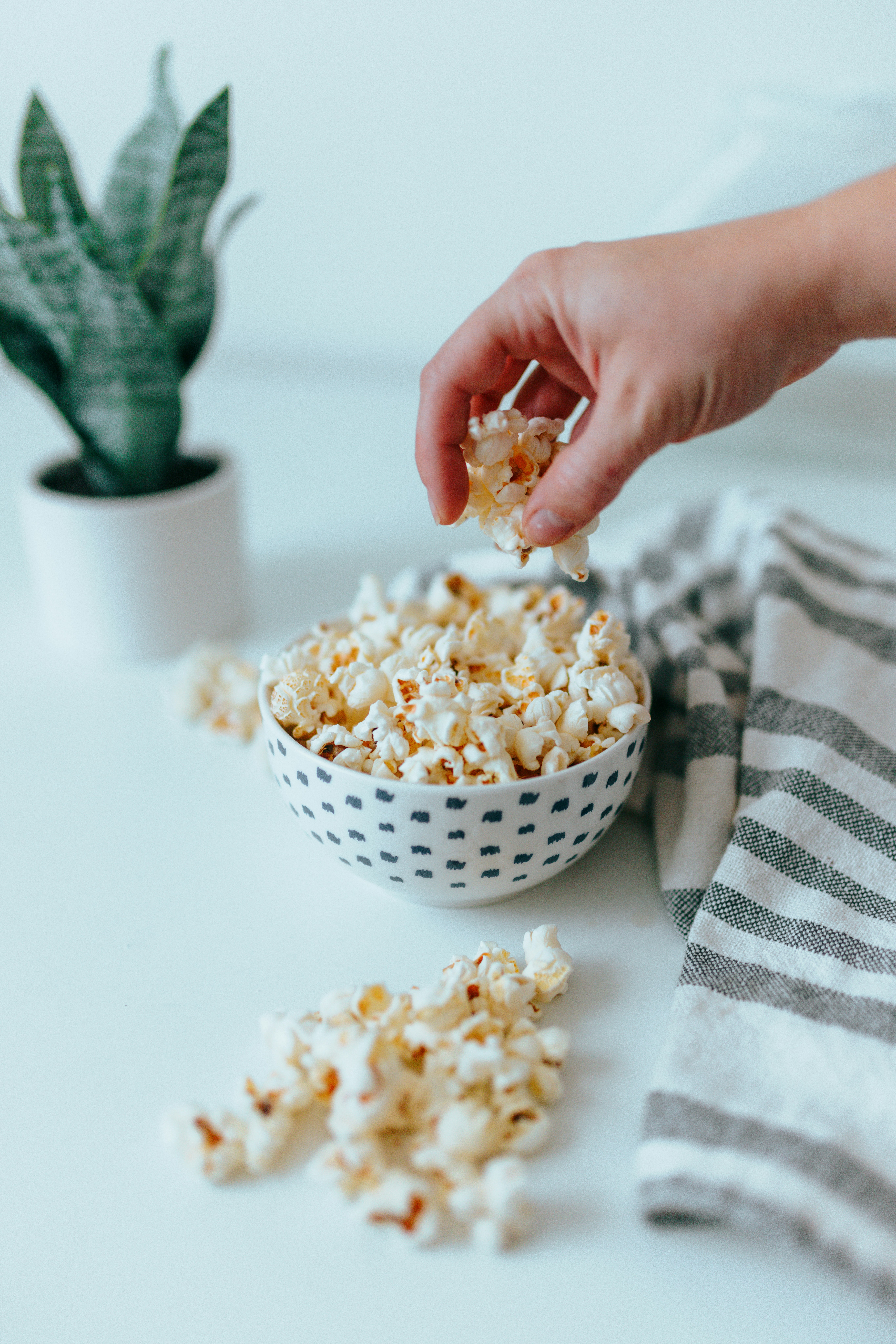 A hand reaches for popcorn from a patterned bowl on a white surface, with scattered popcorn, a potted plant, and a striped cloth nearby.