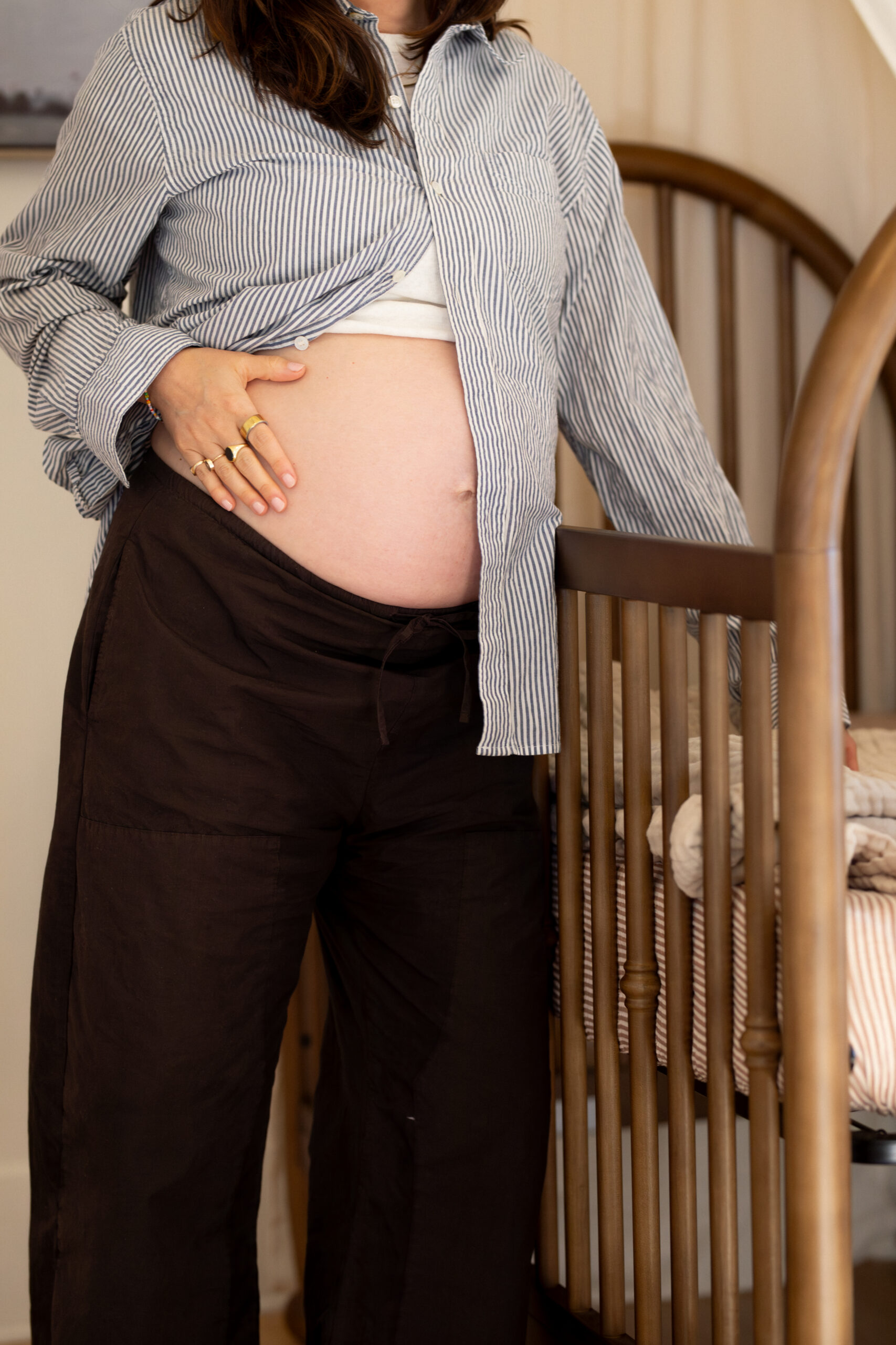 A pregnant person stands beside a wooden crib, one hand resting on their exposed belly and the other holding the crib rail.