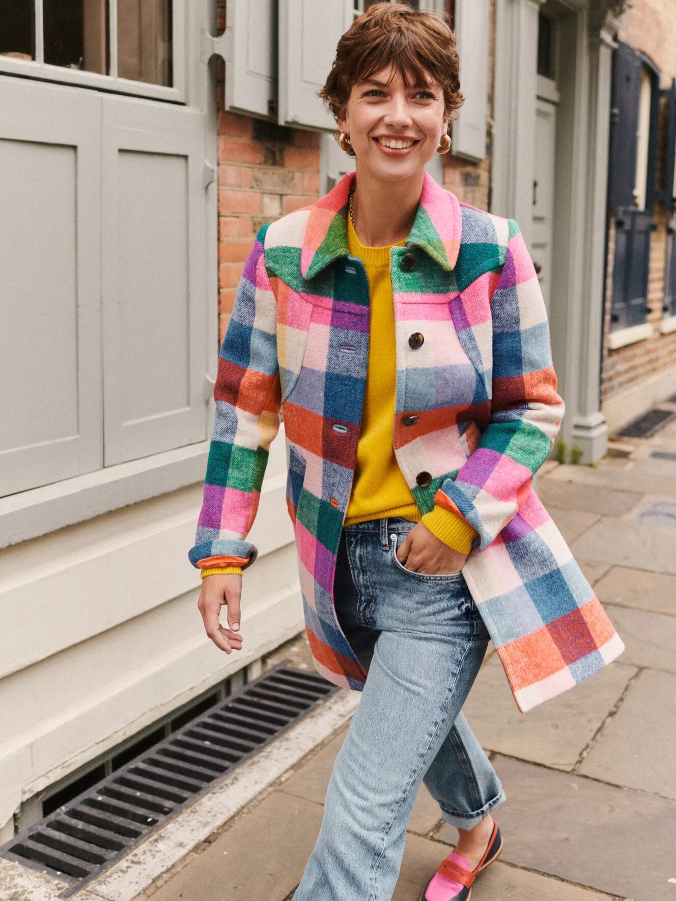 A model walking o a street, smiling, wearing a Boden colorful checkered button down coat. 