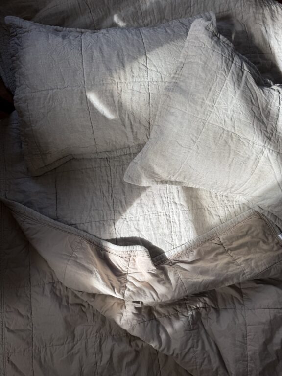 A crumpled gray bedspread and two pillows are illuminated by soft sunlight in a bedroom.