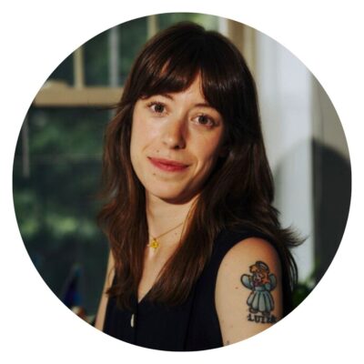 A person with long brown hair and a visible tattoo on their upper arm is indoors, lounging comfortably on an organic mattress while looking at the camera.