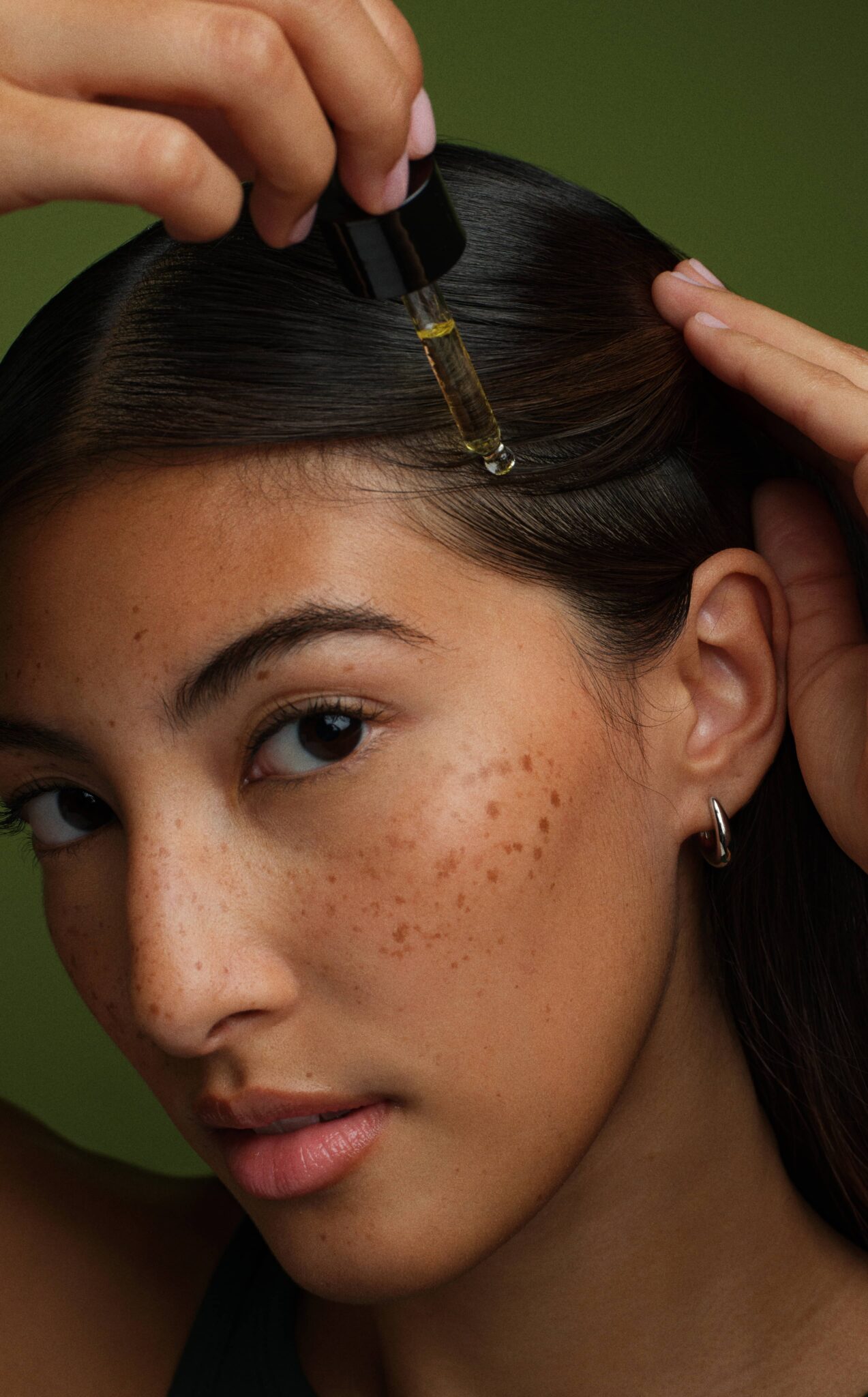 A person applies oil to their scalp with a dropper. The person has long dark hair, hoop earrings, and visible freckles. The background is green.