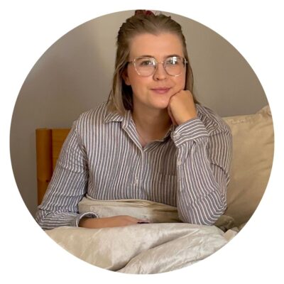 A person with glasses and a striped shirt is sitting on a bed, thoughtfully resting an arm on their chin while contemplating an upcoming mattress review.