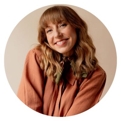 Person with wavy, shoulder-length hair wearing a rust-colored top and a green scarf, smiling at the camera like they've just found the perfect organic mattress.
