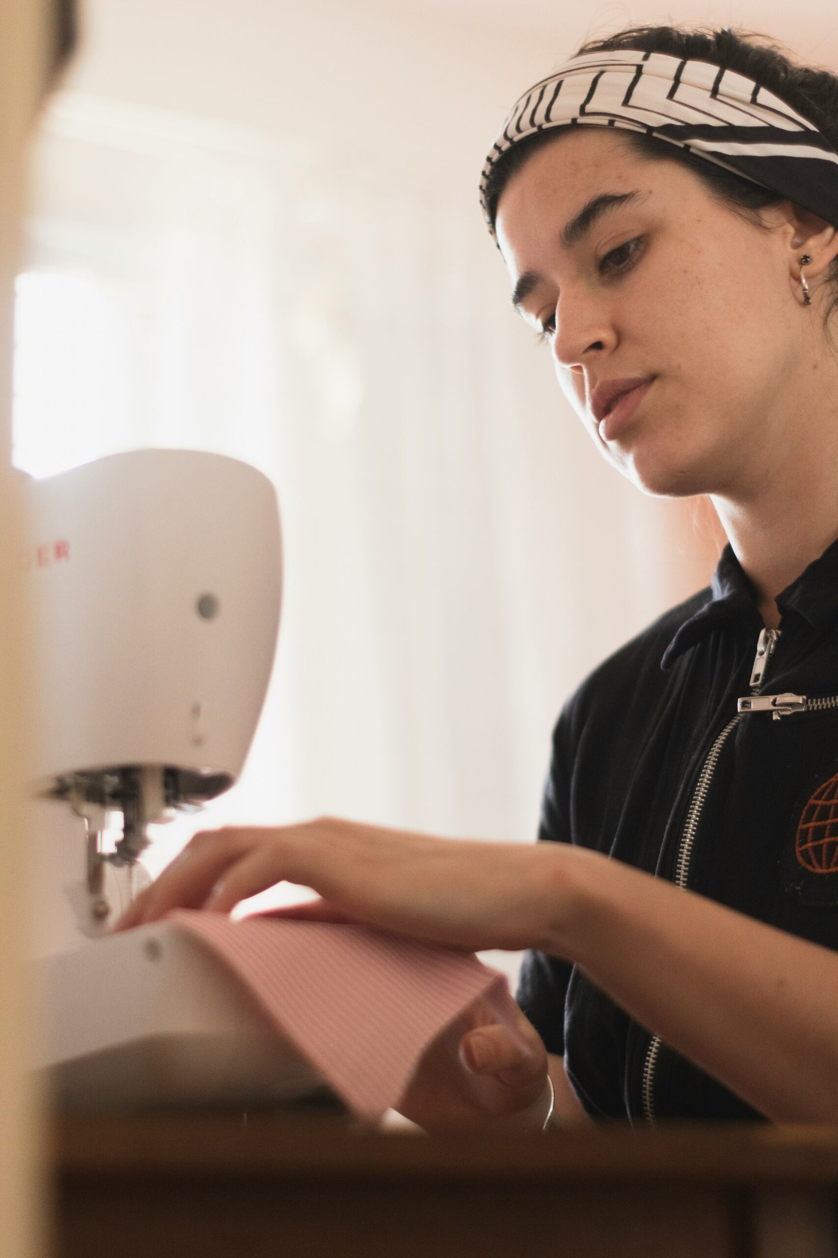 A woman wearing a patterned headband uses a sewing machine to sew a piece of pink fabric.