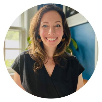 A smiling woman with long brown hair wearing a black top stands in front of a blue wall, adding charm to the serene setting. Behind her, a lush plant hints at nature's beauty—just like choosing an organic mattress for a healthier night's sleep.