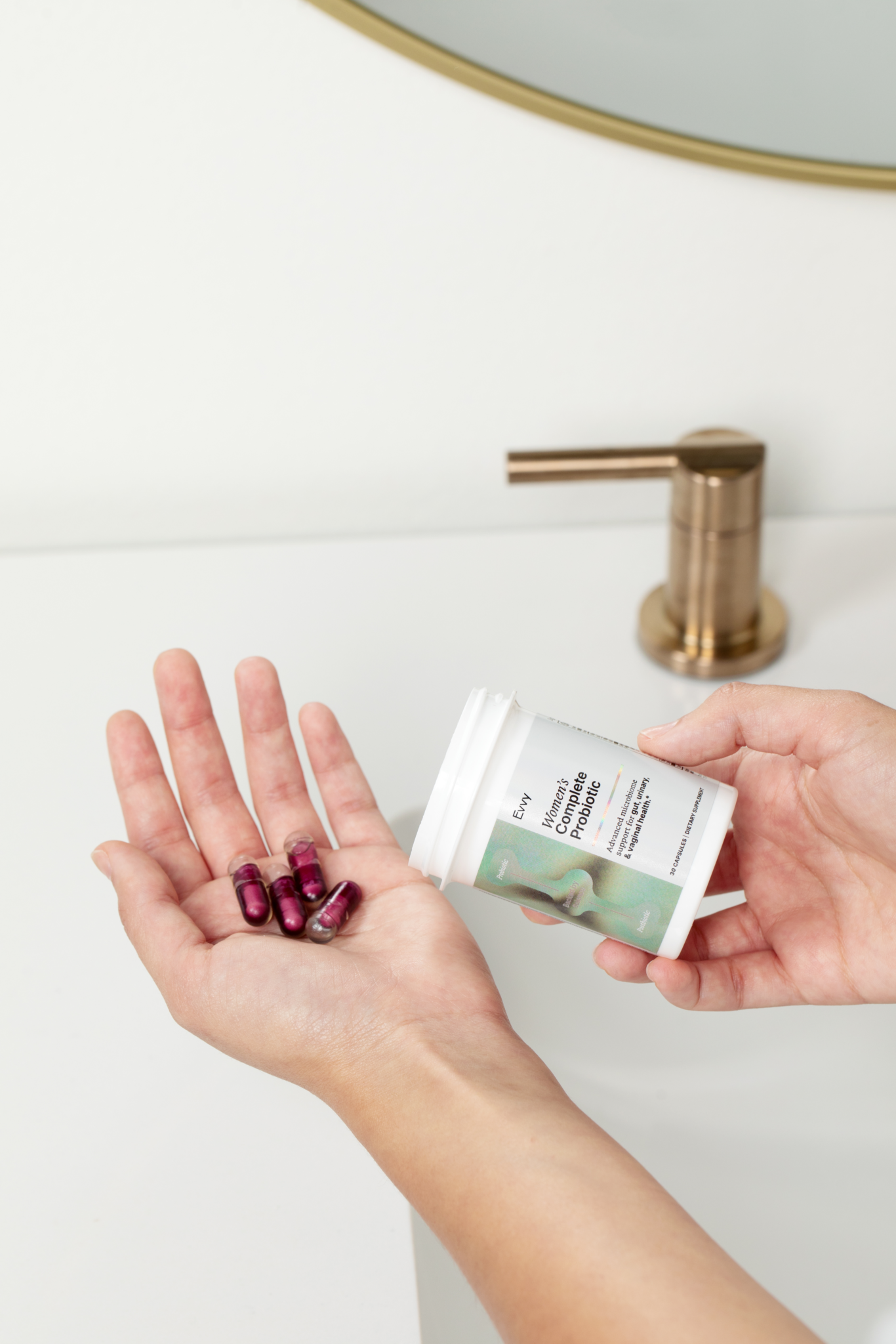 A person holds several purple capsules in one hand and a white pill bottle in the other over a white bathroom sink with a gold faucet.