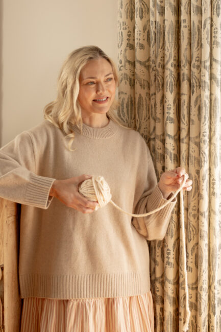 A woman with blonde hair in a beige sweater holds a ball of yarn and stands beside a patterned curtain, looking slightly to the side and smiling.