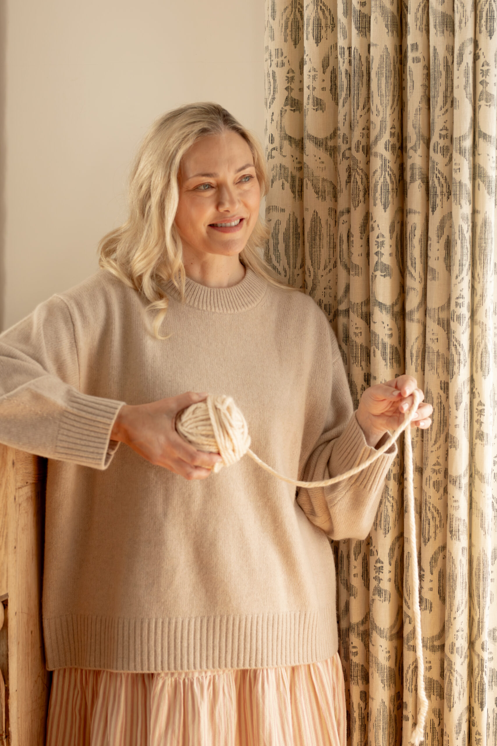 A woman with blonde hair in a beige sweater holds a ball of yarn and stands beside a patterned curtain, looking slightly to the side and smiling.