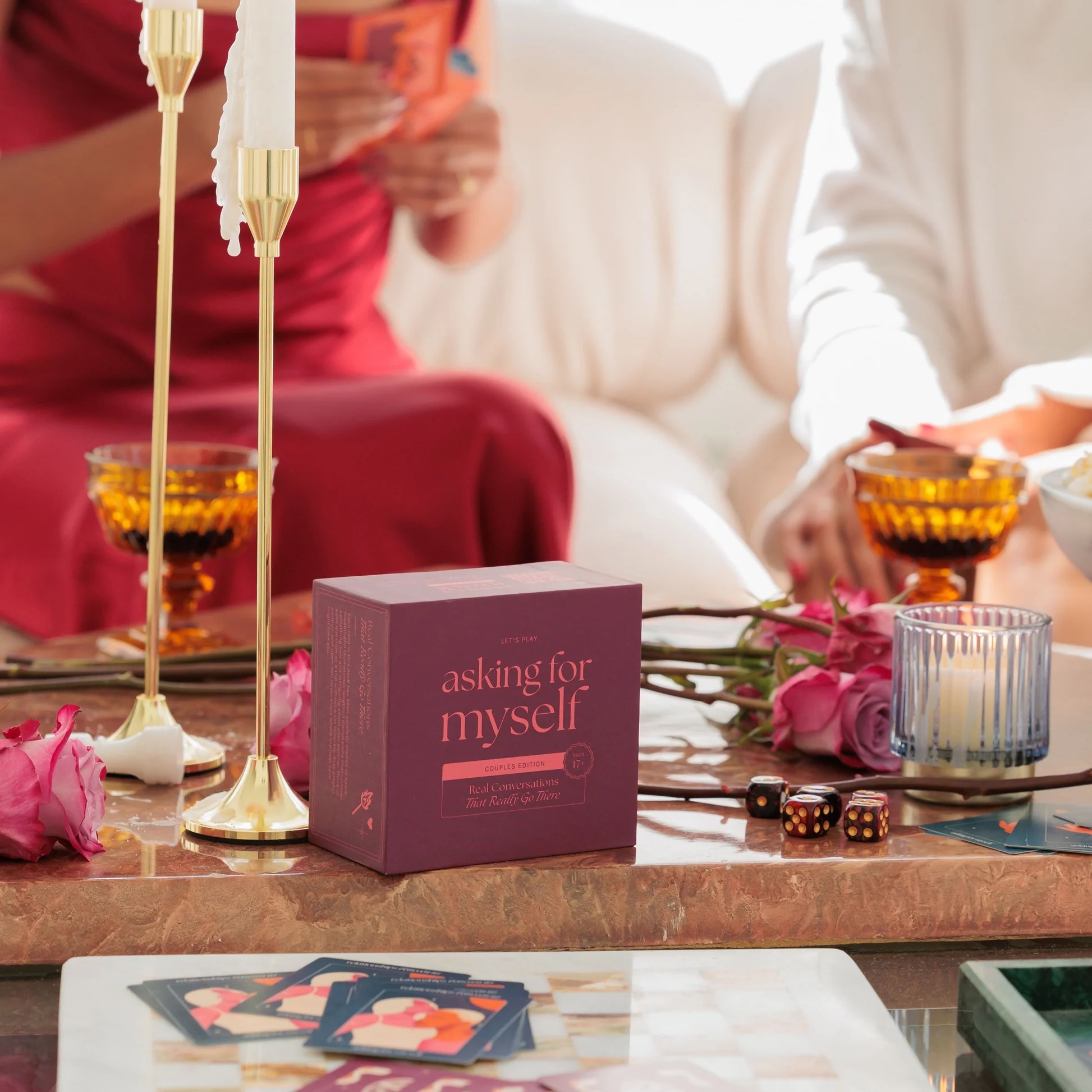 A maroon box labeled "asking for myself" sits on a table with candles, roses, cards, and glassware. Two people are seated nearby, partially visible.