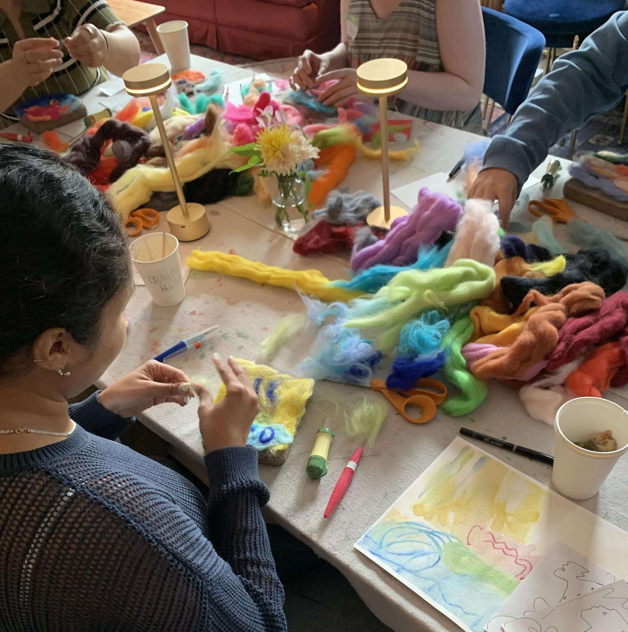 People sit around a table working on a needle felting project, with colorful wool, tools, and art supplies spread across the surface.
