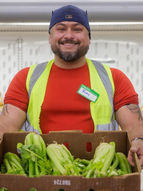 A man wearing a high-visibility vest and a backwards cap holds a box of leafy green vegetables, smiling at the camera. He has a name tag that reads “Rick.”.