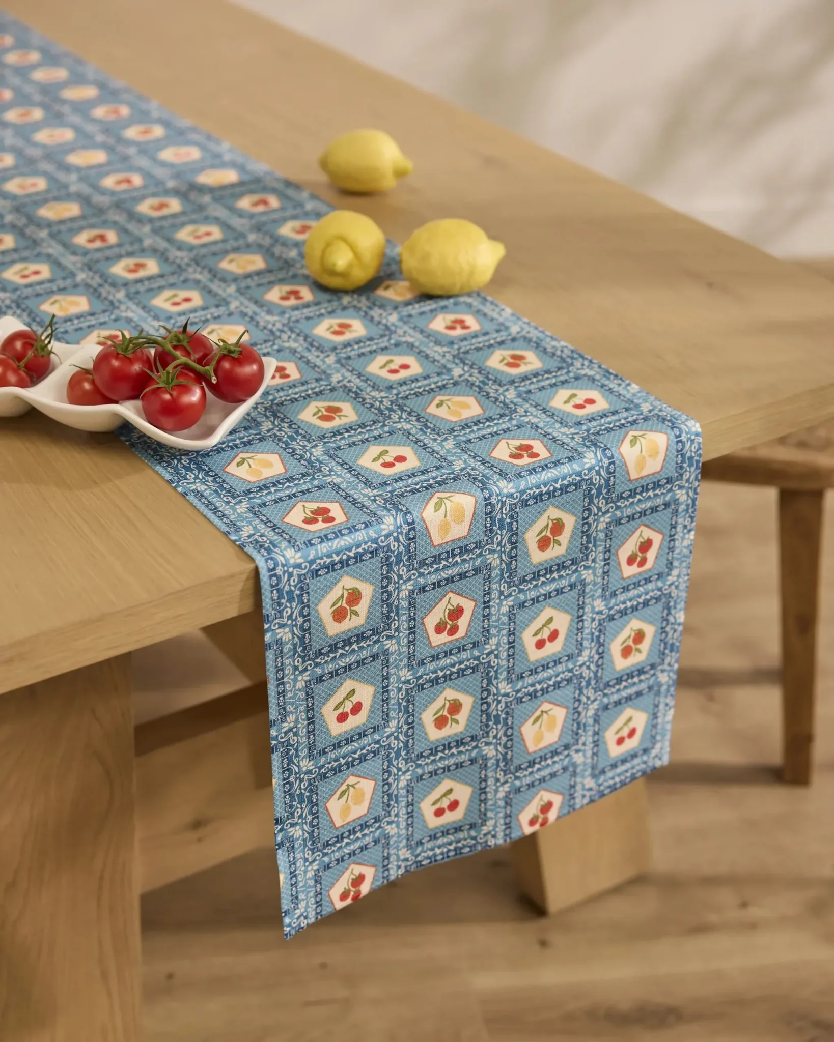 A light wooden table with a blue patterned table runner, three lemons, and a ceramic dish holding vine tomatoes.