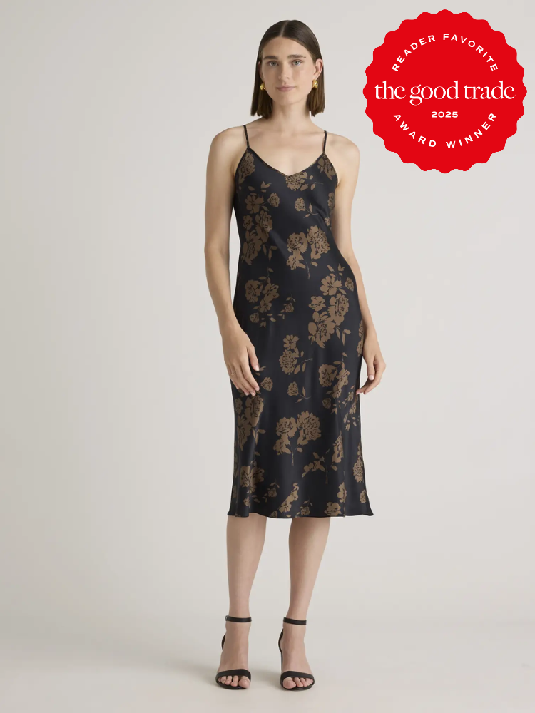 A person models a black sleeveless dress with a brown floral pattern. They stand with arms relaxed by their sides. A red badge on the right reads "the good trade 2025 reader favorite award winner.