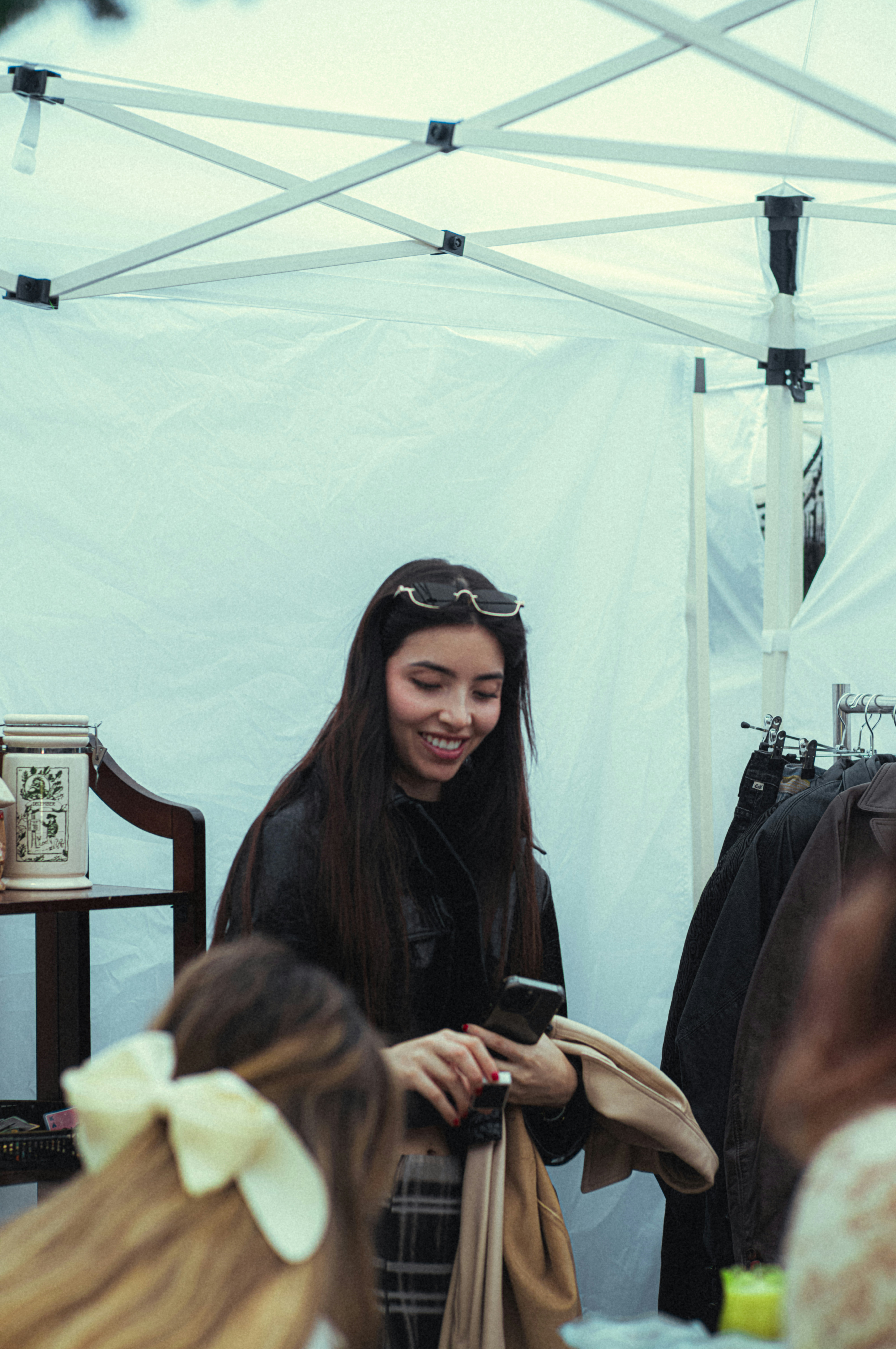 A woman with long hair stands under a white tent, smiling while holding a phone and coat. Coats hang nearby and another person with a hair bow is in the foreground.