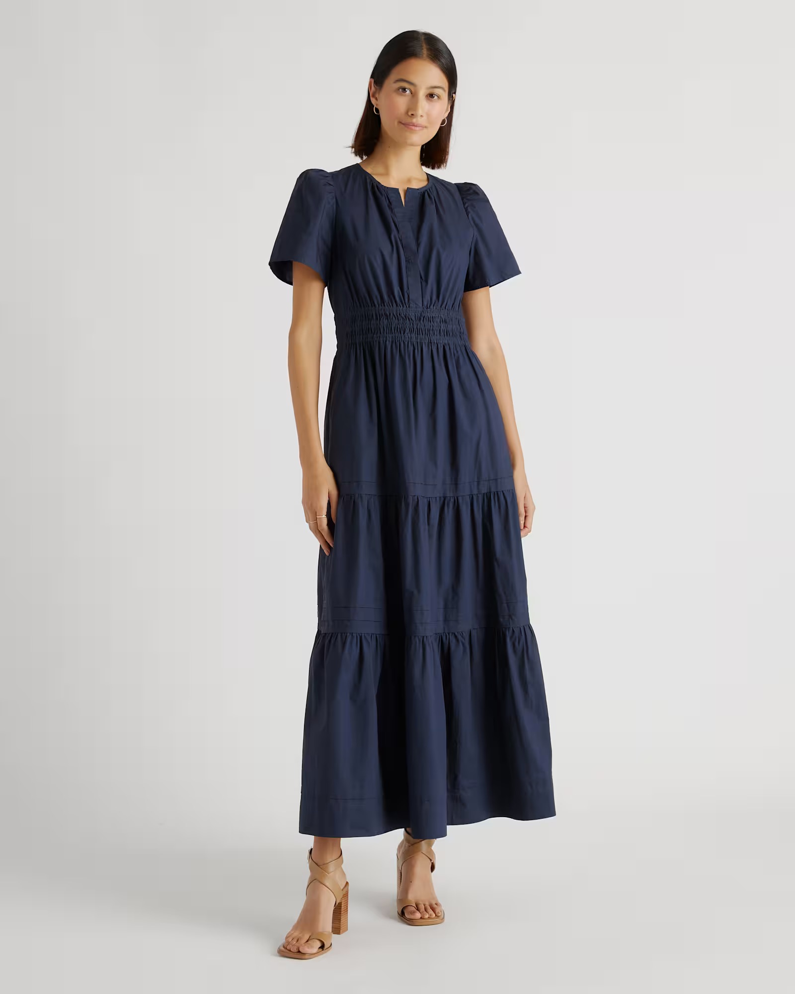A woman stands wearing a navy blue, short-sleeve, ankle-length dress with a fitted waist and tiered skirt, paired with tan heeled sandals.