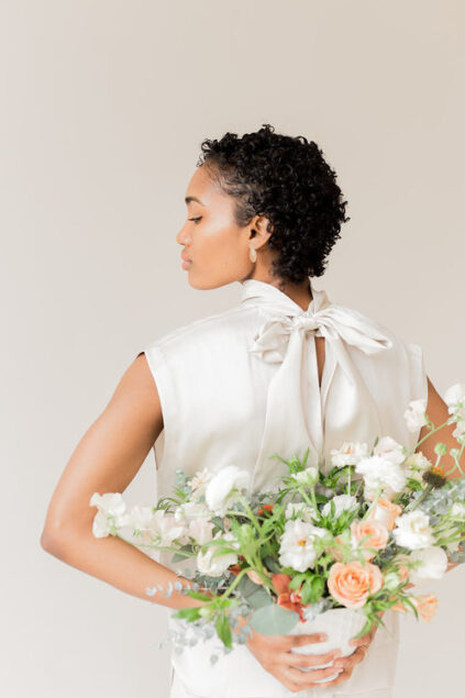 A person with short curly hair, wearing a light-colored top with a back bow, holds a bouquet of white and peach flowers against a neutral background.