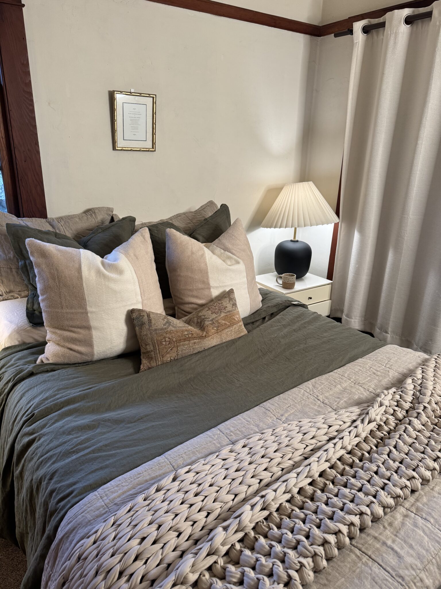 A neatly made bed with green and beige bedding, large decorative pillows, a knit blanket, and a nightstand with a lamp and a small black object beside cream curtains.