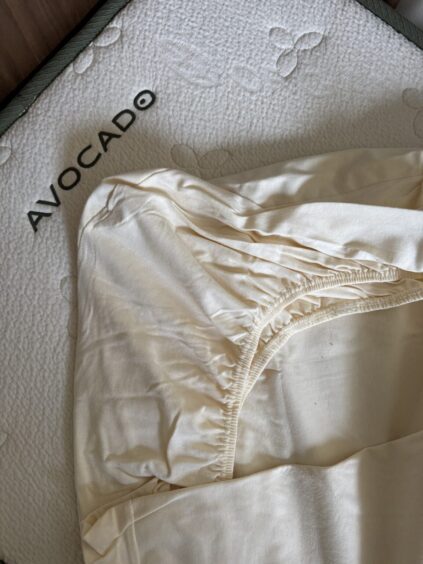 A close-up of a cream-colored fitted sheet partially covering an Avocado brand mattress with a textured surface.