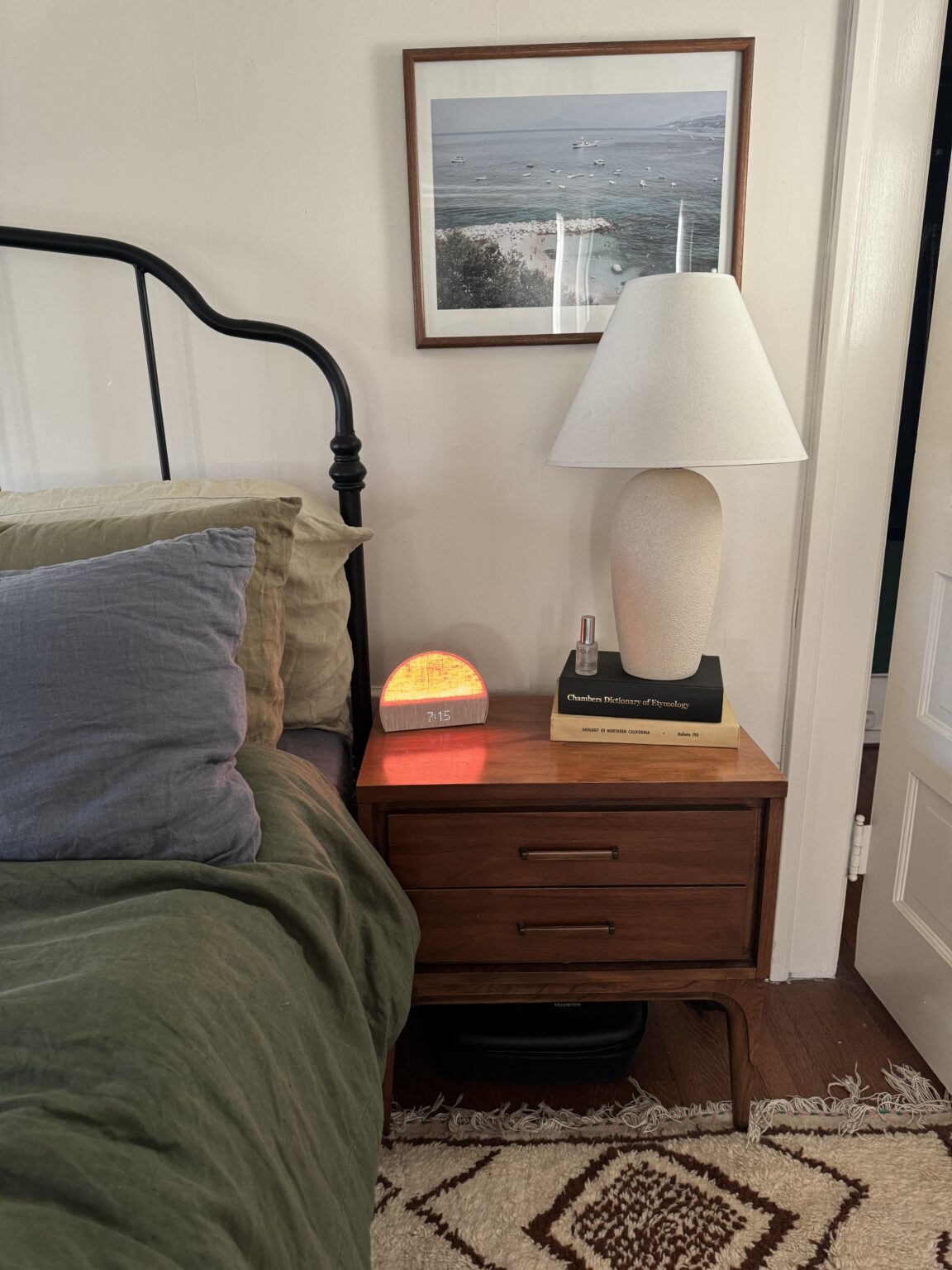 A neatly made bed with green bedding is next to a wooden nightstand holding a lamp, a digital clock with an orange glow, two books, and a framed photo on the wall above.