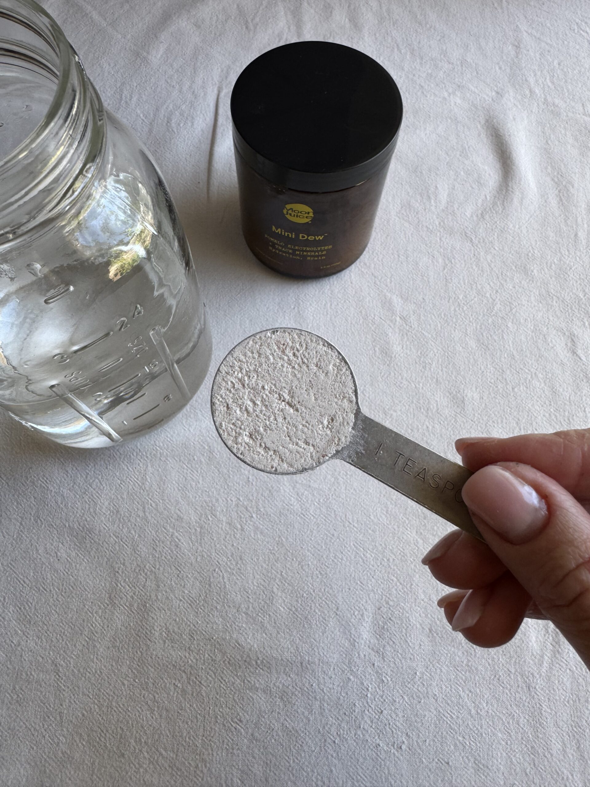 A hand holds a metal teaspoon of white powder over a white surface, next to a glass jar of water and a black-lidded container labeled "Mini Dew.