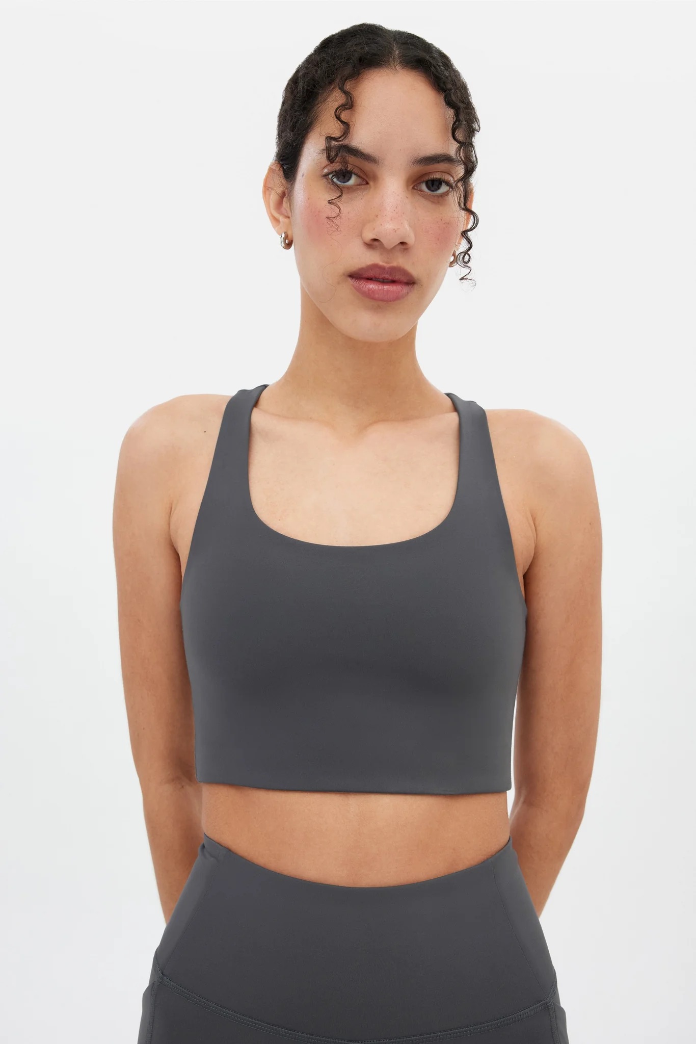 A woman stands against a plain background wearing a dark gray sports bra and matching high-waisted leggings.