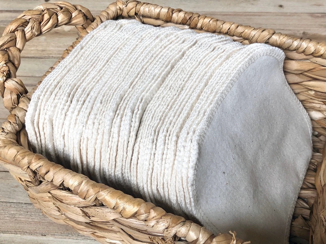 A woven basket filled with a stack of reusable, white cloth wipes or pads, placed on a wooden surface.