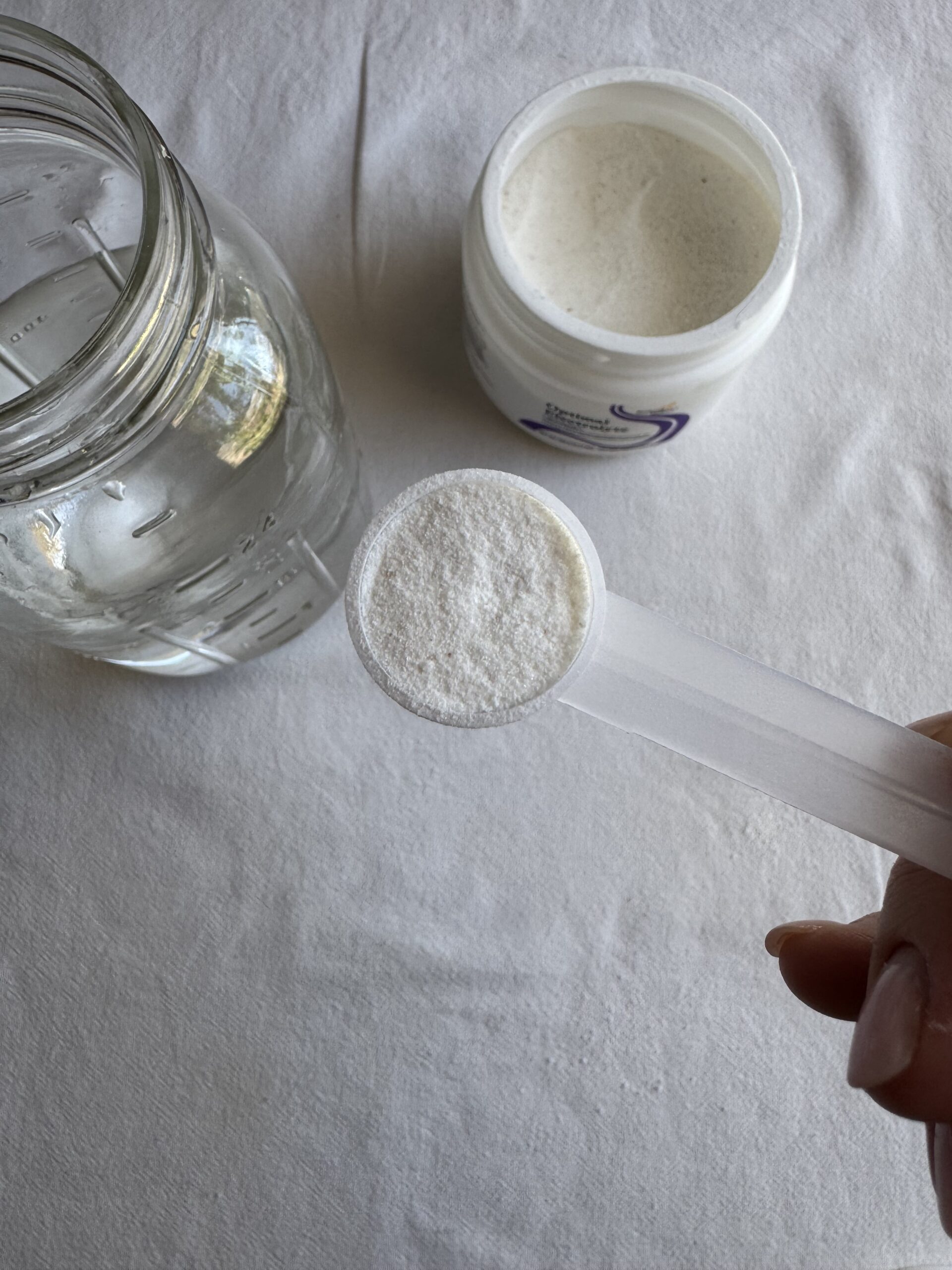A hand holds a plastic scoop of white powder above a glass jar of water and an open container of powdered supplement on a white surface.