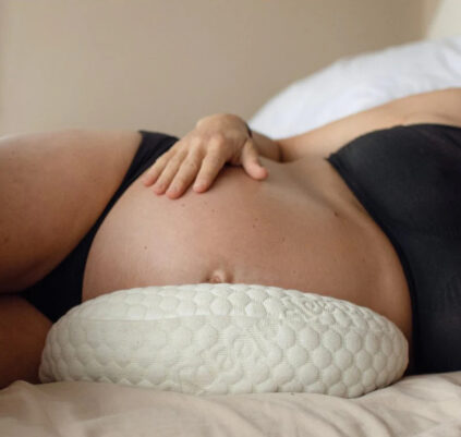 A person lying in bed with a visible pregnant belly, wearing a black bra and using a supportive pillow under their belly.